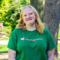 Young woman with a green shirt, black shorts, and blonde hair smiles outside for her picture. Photo captured by Jaden Mezetin Photography.