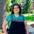 Young woman with brunette hair wearing a green shirt and overalls stands seriously in front of a tree for her photo. Photo captured by Jaden Mezetin Photography.