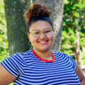 Young woman with curly brunette hair, wearing a striped shirt, glasses, and hoop earrings smiles for her photo. Photo captured by Jaden Mezetin Photography.