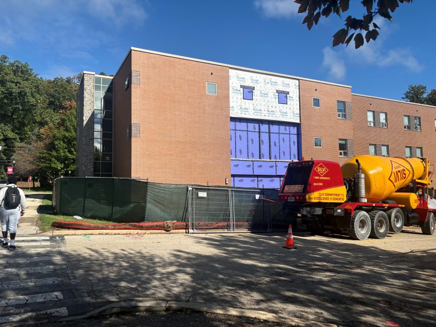 Centennial Hall’s brick exterior with a section of the front wall stripped to blue waterproofing, a green construction fence, and a concrete mixer truck parked in front.